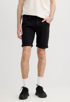 Hombre que lleva pantalones cortos negros remangados, calcetines blancos, zapatillas negras y una camiseta blanca metida por dentro, de pie frente a un fondo liso.