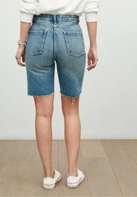 Shorts in denim chiaro con orlo grezzo, vita alta e due tasche posteriori. Indossati con sneakers bianche su un pavimento in legno.