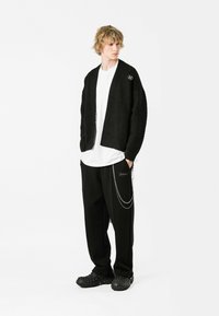 Cardigan negro con textura, camisa blanca, pantalones negros holgados con detalles de cadena plateada y zuecos negros. Silueta general relajada y esquema de color monocromático.