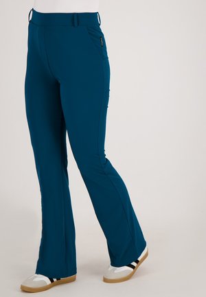 Persoon in teal kleurige flared pants met zakken en witte sneakers met zwarte strepen, staande op een lichte achtergrond.