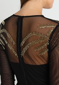 Haut noir transparent à manches longues avec broderie de perles tubulaires dorées en motifs de feuilles sur le haut du dos et les épaules, avec une fermeture éclair centrale.