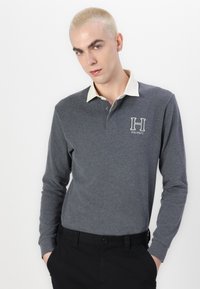 Šedé polo tričko s dlhým rukávom a krémovým golierom, s vyšitým logom "H" a gombíkovým zapínaním. Nosené s čiernymi nohavicami.
