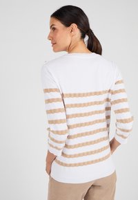 Witte trui met beige golvende strepen, driekwartmouwen en een geribbelde zoom. Zachte textuur met knoopdetail op de schouder.