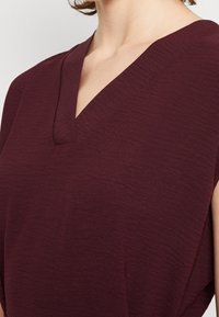 Haut bordeaux texturé avec un décolleté en V et des manches courtes, mettant en avant un tissu doux et léger ainsi qu'une profondeur visuelle subtile.