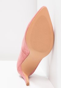Chaussure à talon haut en daim rose à bout pointu, vue de la semelle avec un motif ondulé, sur fond blanc.