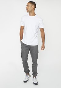 Witte t-shirt met ronde hals, grijze cargobroek met zijzakken, en hardloopschoenen met blauwe en witte accenten, staand tegen een effen achtergrond.