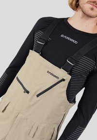 Pantaloni salopette impermeabili beige con spalline nere regolabili, dotati di due tasche con zip e pieghe per una maggiore flessibilità, abbinati a un sottogiacca a maniche lunghe nera.