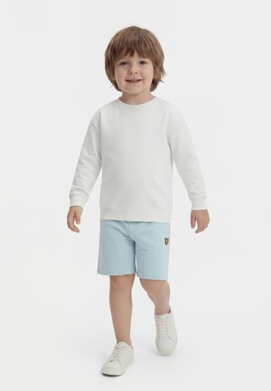 Nuori poika vaaleanruskein hiuksin kävelee, hänellä on yllään valkoinen pitkähiha- paita, vaaleansiniset shortsit, joissa on pieni keltainen logo, ja valkoiset tennarit.