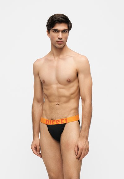 Jeune homme aux cheveux courts et foncés portant un slip noir avec une ceinture orange étiquetée "Diesel" sur un fond clair uni.