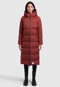 Lang maroon pufferjakke med hætte, med lynlås fortil, to sidelommer med lynlås og sorte accenter. Bæres med sorte bukser og ankelstøvler.