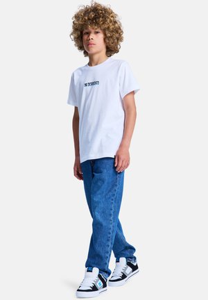 Jongere met krullend haar, gekleed in een wit T-shirt met grafisch ontwerp, blauwe spijkerbroek en zwart-witte sneakers, staand tegen een effen achtergrond.