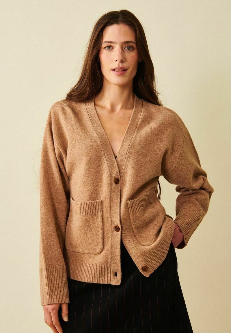 Pitkähiuksinen nainen, jolla on päällään beige neulejakku, jossa on napit ja etutaskut, sekä musta, hienovaraiset pystyraidat sisältävä hame.