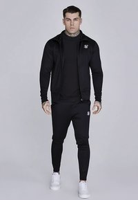 Completo sportivo nero composto da una giacca con zip e pantaloni aderenti, entrambi con una texture liscia e un piccolo dettaglio logo. Scarpe da ginnastica bianche completano il look.