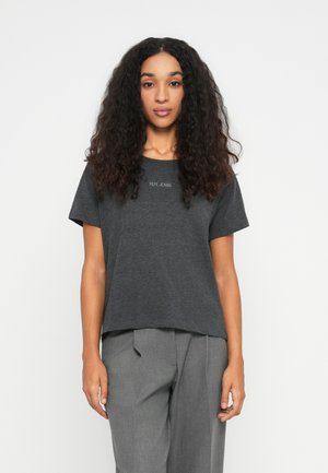 Kvinde med langt, krøllet hår iført en mørkegrå "Pepe Jeans" t-shirt og lysegrå plisserede bukser, stående mod en simpel hvid baggrund.