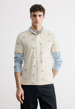 Giovane uomo che indossa una polo beige con stampe nautiche sopra una camicia a maniche lunghe azzurro chiaro e jeans scuri, in piedi contro uno sfondo bianco.