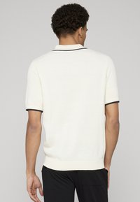 Polo en maille à manches courtes couleur crème avec bordures bleu marine au col et aux poignets, présentant un tissage texturé et un design uni.