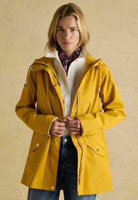 Geltonas vandeniui atsparus džemperis su gobtuvu, priekiniu užtrauktuku, raišteliu ir dviem įstrižais kišenėmis. Sujungtas su balta marškinėliais ir raštuota šaliku.