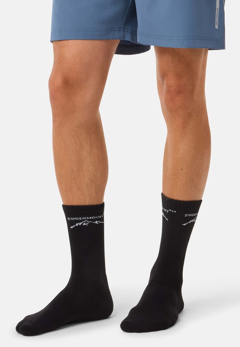 Nogi w czarnych skarpetkach do połowy łydki z białym logo "Swedemount" i górą, w połączeniu z niebieskimi szortami na jednolitym białym tle.