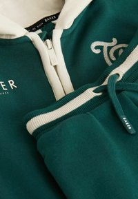 Sudadera con cremallera verde que presenta una capucha beige, puños acanalados, logotipo bordado y detalles de cordón, hecha de una suave tela texturizada.