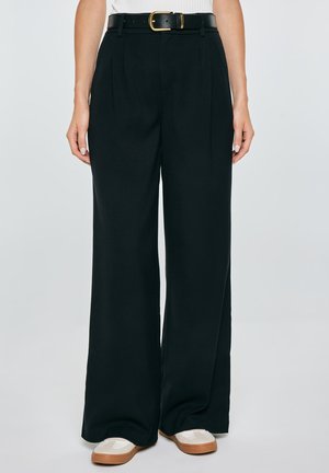 Kobieta nosząca czarne, wysokowciskane spodnie z szerokimi nogawkami i bocznymi kieszeniami, czarny pasek ze złotą klamrą oraz białe buty z brązowymi podeszwami.