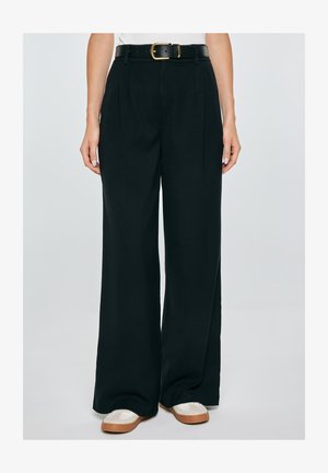 Kobieta nosząca czarne, wysokowciskane spodnie z szerokimi nogawkami i bocznymi kieszeniami, czarny pasek ze złotą klamrą oraz białe buty z brązowymi podeszwami.