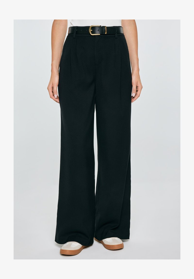 Naine, kes kannab kõrge vöökohaga musta laia lõikega pükse küljetaskutega, musta vööd kuldse pandlaga ja valgeid jalatseid pruunide talladega.