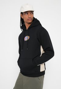 Sudadera negra con bolsillo frontal, que presenta un logo colorido y acentos crema a los lados. El material parece suave, con un corte relajado y capucha con cordón.