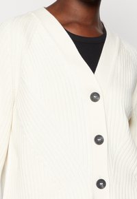 Krämfärgad kofta med ribbad textur, V-ringad design och tre mörka knappar. Bärs över en svart skjorta, vilket framhäver lager på lager-stilen.