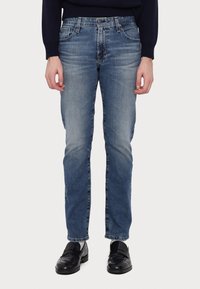 Henkilö, joka on pukeutunut slim-fit sinisiin farkkuihin, mustiin loafer-kenkiin, valkoisiin sukkiin ja tummanväriseen pitkähihaiseen paitaan, seisoo yksinkertaista valkoista taustaa vasten.