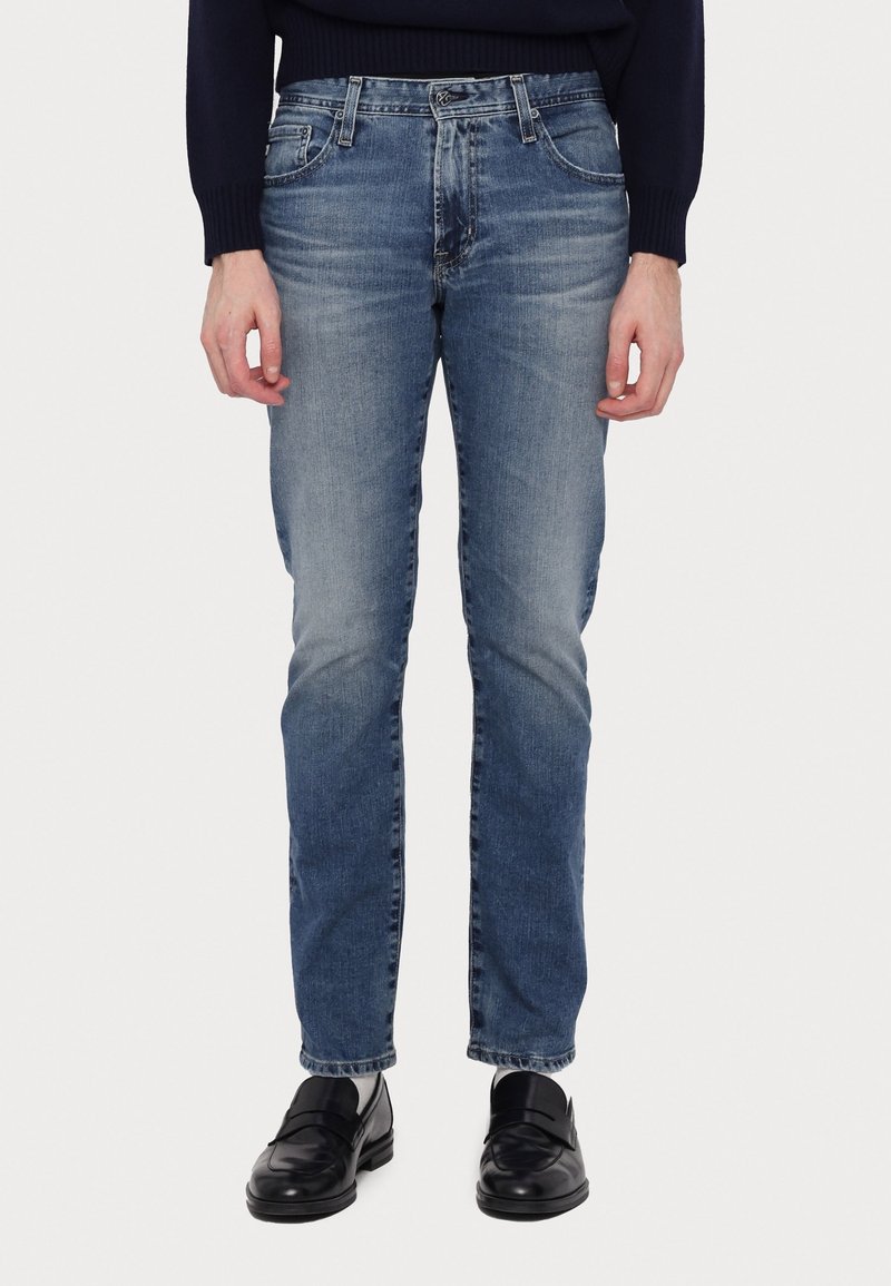 Henkilö, joka on pukeutunut slim-fit sinisiin farkkuihin, mustiin loafer-kenkiin, valkoisiin sukkiin ja tummanväriseen pitkähihaiseen paitaan, seisoo yksinkertaista valkoista taustaa vasten.