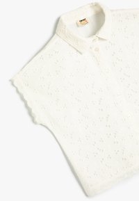 Hvid kortærmet skjorte lavet af struktureret stof med broderede blomsterdesigns, bølgede kanter og knaplukninger.