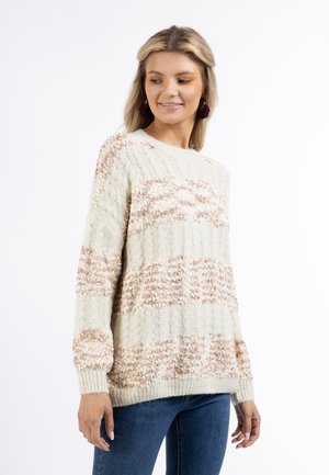 Donna che indossa un maglione lavorato a maglia crema e marrone chiaro con maniche lunghe, abbinato a jeans blu, sorride leggermente e guarda verso il basso.