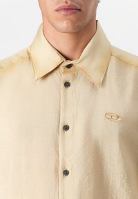Camisa beige de botones con tejido texturizado, que presenta un cuello con puntas y botones oscuros. Incluye un pequeño logotipo bordado en el pecho.