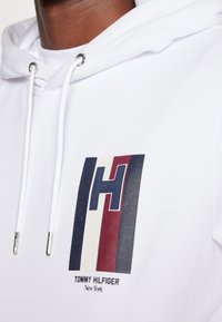 Felpa bianca con un grafico che presenta strisce rosse, blu navy e crema, e un audace logo "H". Gli occhielli in argento adornano il cordino.