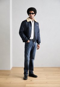 Cazadora bomber azul marino con un collar de piel sintética beige, camisa blanca, vaqueros azules y zapatos negros. Diseño sencillo, sin patrones ni logotipos visibles.