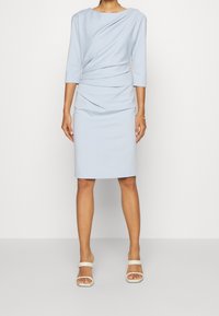 Vestido ajustado de un azul claro en tela elástica, con un escote barco y mangas tres cuartos, y detalles fruncidos en la cintura.