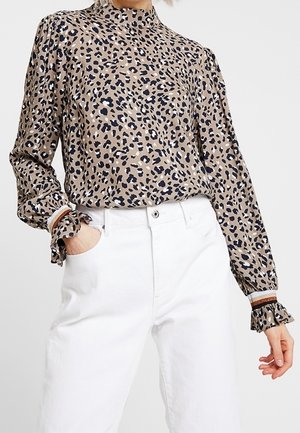 Femme portant un chemisier à manches longues imprimé léopard avec des poignets à volants et un col montant, associé à un pantalon blanc taille haute.