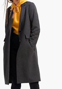 Abrigo gris de mezcla de lana con un ajuste entallado, diseño de un solo botón, bolsillos con solapa y solapas con muesca, combinado con una sudadera con capucha amarilla debajo.