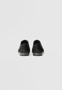 Chaussures en cuir noir avec une texture lisse, arrière arrondi et semelle en caoutchouc grise. Design minimaliste sans lacets ni ornements visibles.