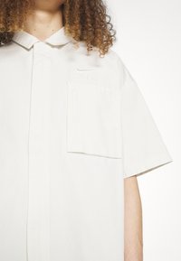 Světle zbarvená košile s krátkým rukávem a límcem, se zesílenou přední kapsou a embosovaným logem "AIR". Měkká tkanina a minimalistický design.