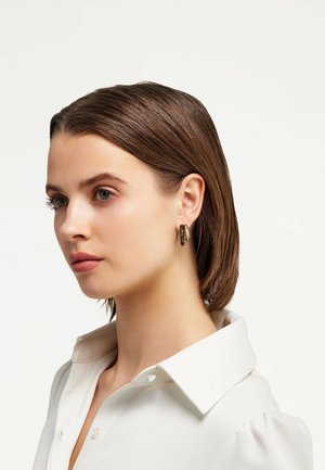 Gold hoop earrings with a textured surface, featuring a shiny finish and a circular design, shown worn by a model with a white collared shirt.