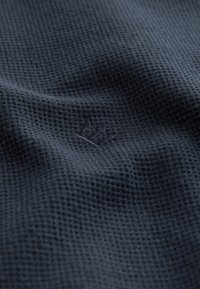 Tessuto strutturato blu navy scuro con un motivo a griglia, caratterizzato da un logo ricamato sottile. Il materiale sembra traspirante e leggero.
