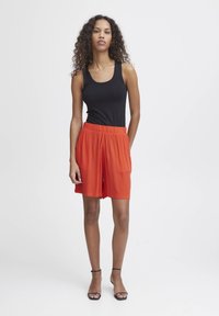 Zwarte mouwloze top gecombineerd met oranje geplooide shorts, met een elastische tailleband. Model staat in zwarte sandalen met bandjes tegen een lichte achtergrond.
