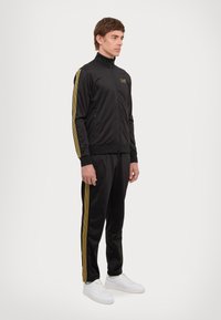 Black tracksuit with gold stripes, zippered jacket, and slim-fit trousers, made of smooth synthetic material. White sneakers complete the look.