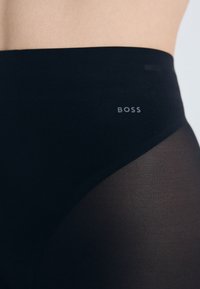 Crne tajice visokog struka sa prozirnim mrežastim panelima. Imaju glatku teksturu i mali srebrni logotip "BOSS" na pojasu.