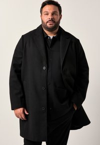 Cappotto in misto lana nero con colletto classico, design a due bottoni e tasche laterali. Indossato sopra una camicia nera.