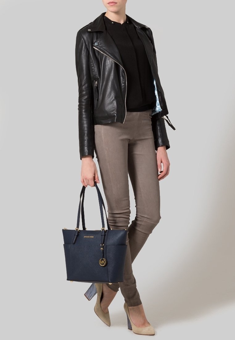 Personne portant une veste en cuir noire, un haut noir, un pantalon taupe, des talons beiges, tenant un sac à main Michael Kors bleu marine avec des accents dorés.