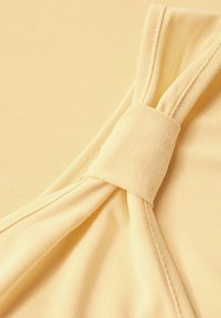 Ljust gult tyg med slät textur, med en knutdetalj vid halsen. Enkel design med subtila sömdekorationer.