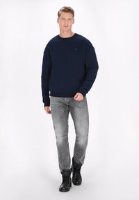 Navy Sweatshirt mit rundem Halsausschnitt, gerippten Bündchen und Saum, getragen mit grauen Jeans und schwarzen ankle boots, mit einem kleinen Anker-Logo auf der Brust.