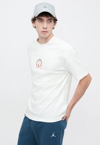 Άσπρο βαμβακερό t-shirt με κοντά μανίκια, με κόκκινο και μαύρο γραφικό σχέδιο στο στήθος. Συνδυάζεται με σκούρο μπλε παντελόνι φόρμας.